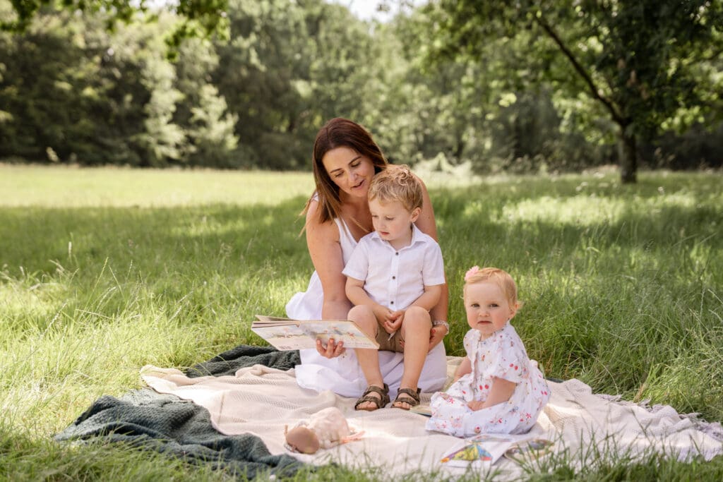 Mum and two children sat on a blanket in the park reading a book
