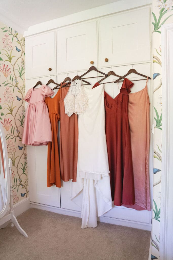 Bridesmaid dresses in rust, terracotta and blush pink hanging next to the wedding dress