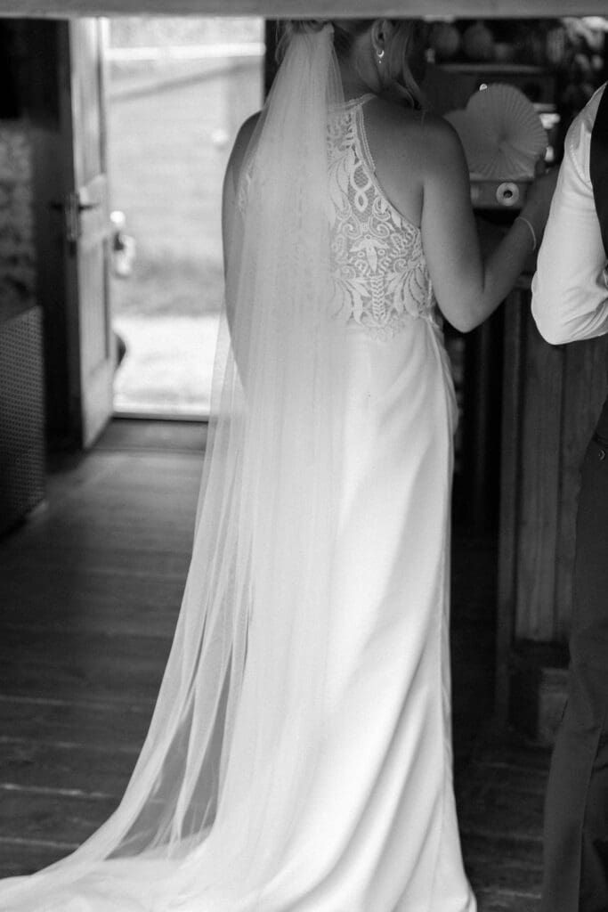Black and white photo of the bride from behind in her long veil, standing in a doorway before the ceremony