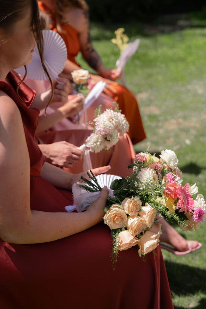 Bridesmaids in rust dresses holding bouquets and paper fans during the outdoor ceremony