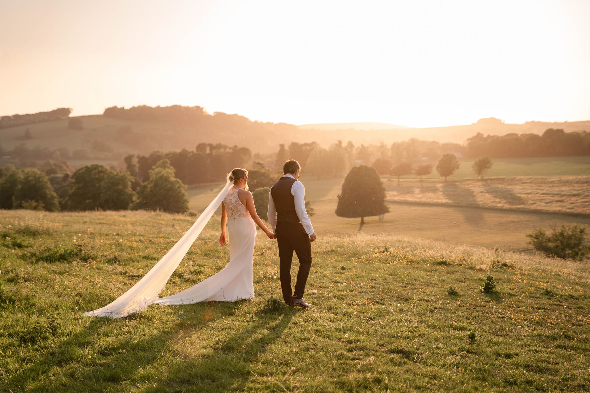 Bride and groom walking hand in hand through the fields at golden hour, her long veil trailing behind, captured by their Cissbury Barns wedding photographer