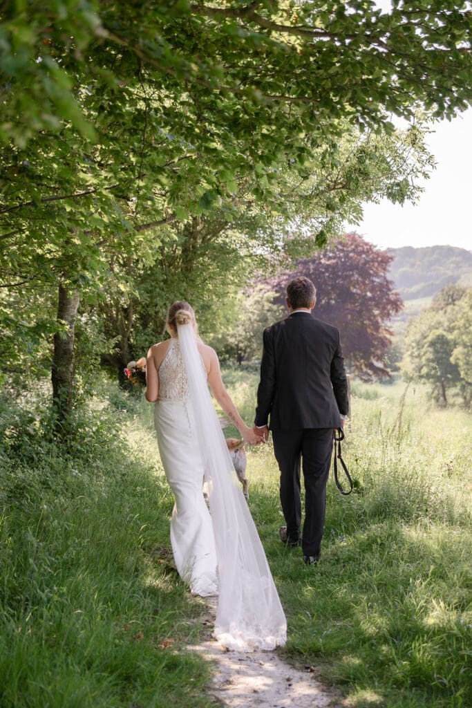 Bride and groom walking along a grassy path through the fields next to the barn with their Labrador