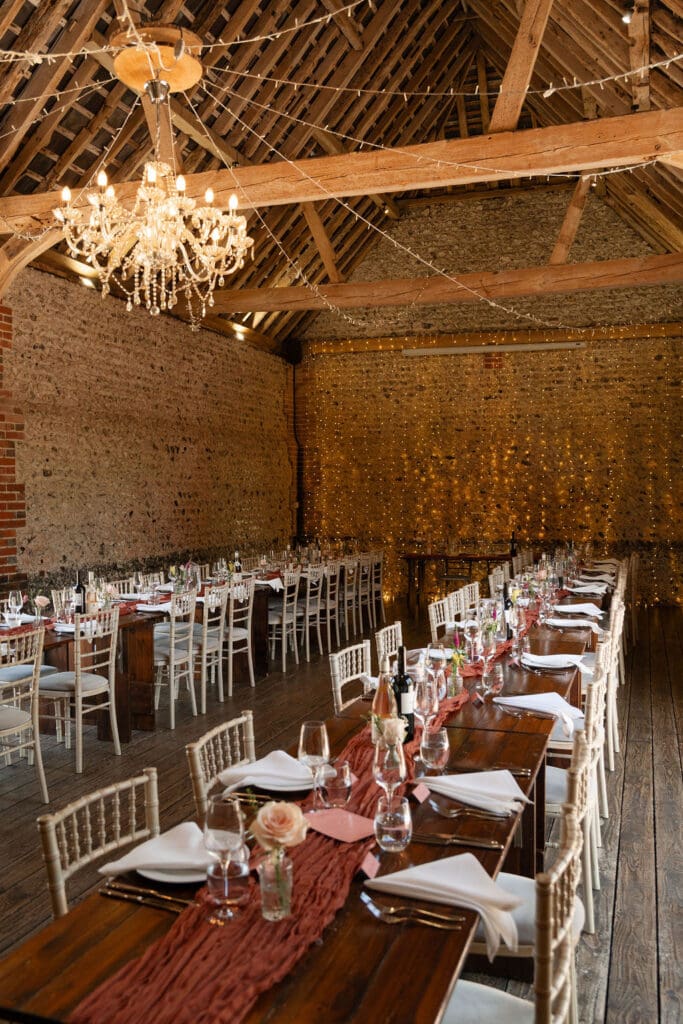 Long tables set for the wedding breakfast inside the barn, with a chandelier and timber beams