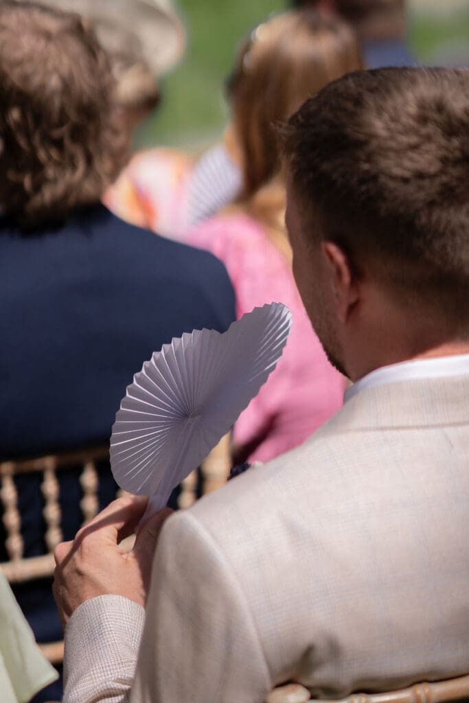 Guests holding paper fans as they watch the outdoor ceremony on a hot May day
